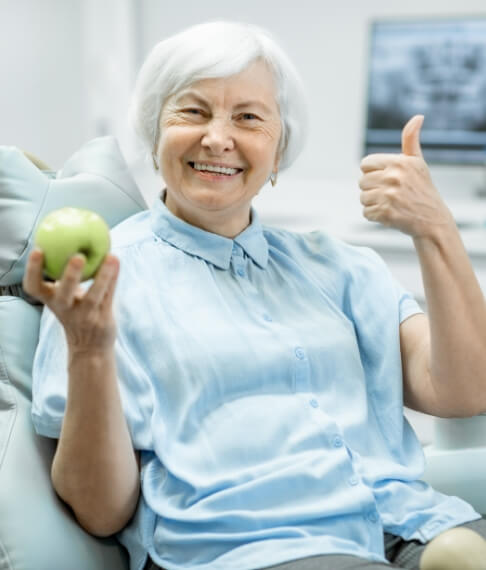 Woman giving thumbs up and holding an apple after dental implant tooth replacement