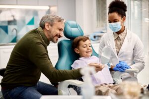 Child in dentist's chair flanked by dentist on one side and father on the other.